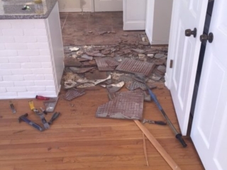 Photo of kitchen tile being removed. Pictures shows broken pieces of tile adjacent to a living room with hardwood flooring.