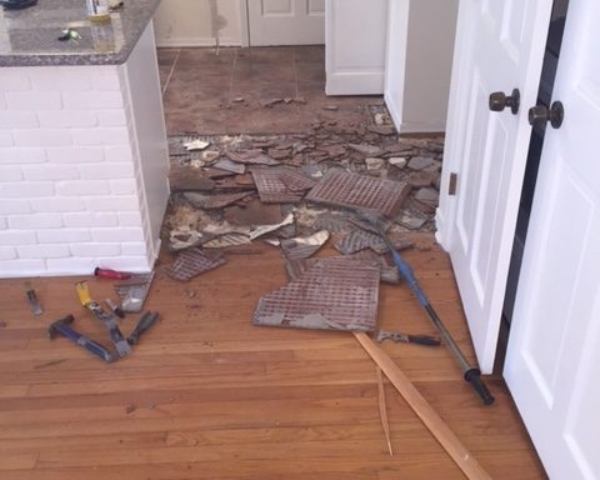 Photo of kitchen tile being removed. Pictures shows broken pieces of tile adjacent to a living room with hardwood flooring.