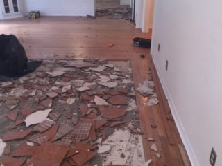 Picture shows broken tile in the process of being removed. It is a square section of a living room surrounded by hardwood flooring.