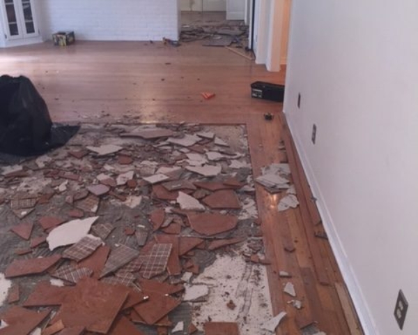 Picture shows broken tile in the process of being removed. It is a square section of a living room surrounded by hardwood flooring.